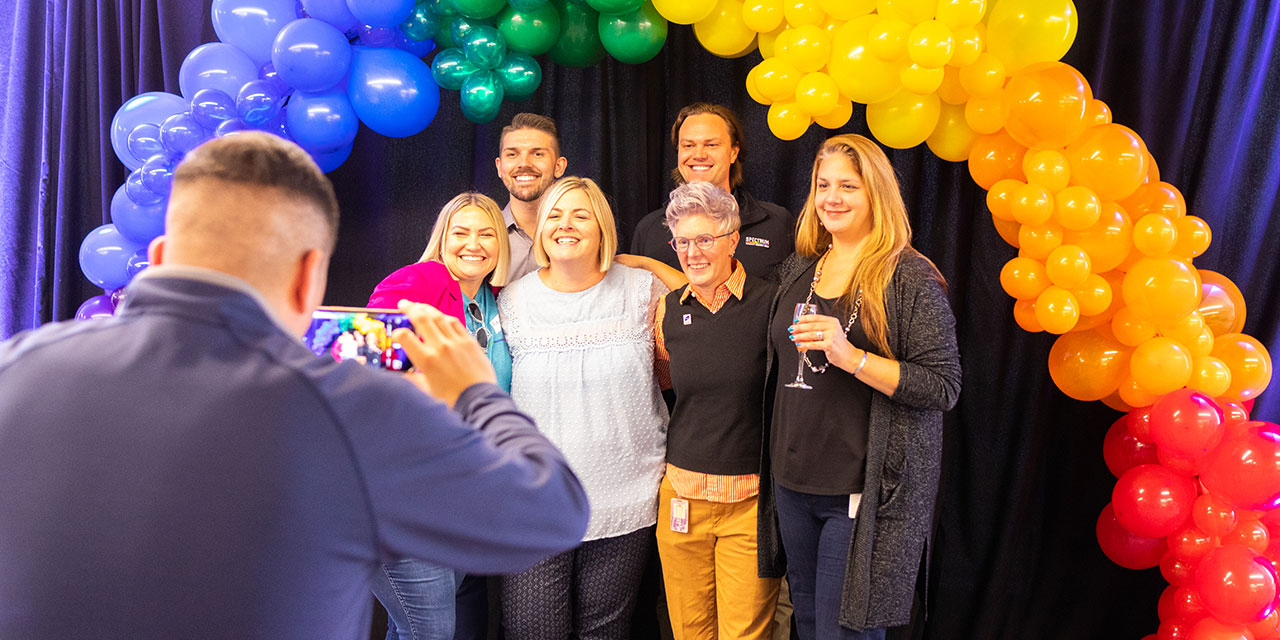 A group of Liberty associates pose for a photo under an arch of rainbow colored balloons at a corporate event