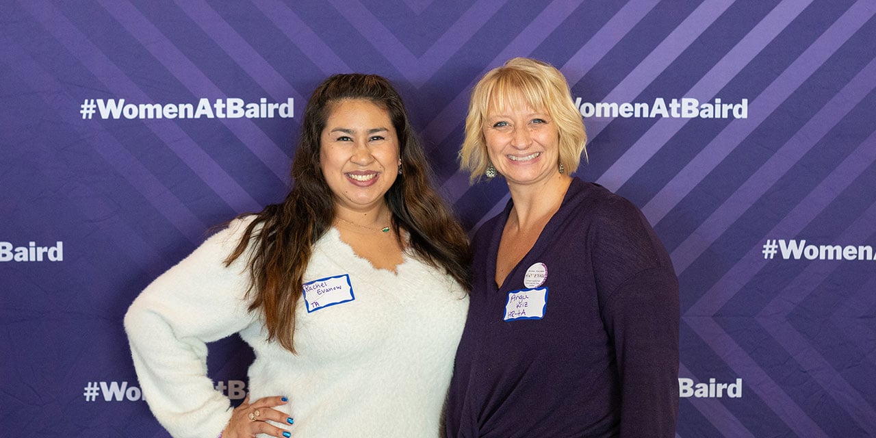 Two Liberty associates in front of a purple banner with #womenatbaird hashtags